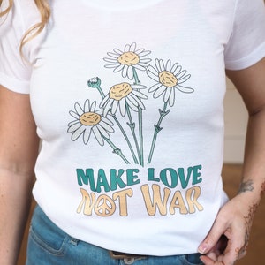 Op de afbeelding: Wit T-shirt met een afbeelding van madeliefjes en de tekst "Make Love Not War" met een vredesteken.