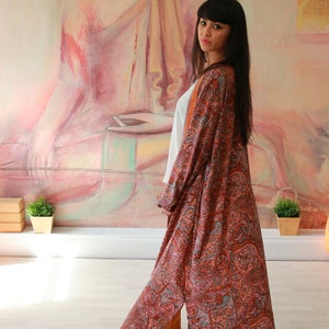 Puede incluir: Una mujer lleva una larga t&uacute;nica de estampado paisley con un top blanco debajo. La t&uacute;nica es una mezcla de colores rojo, naranja y azul. La mujer est&aacute; de pie frente a una pintura de un Buda meditando.