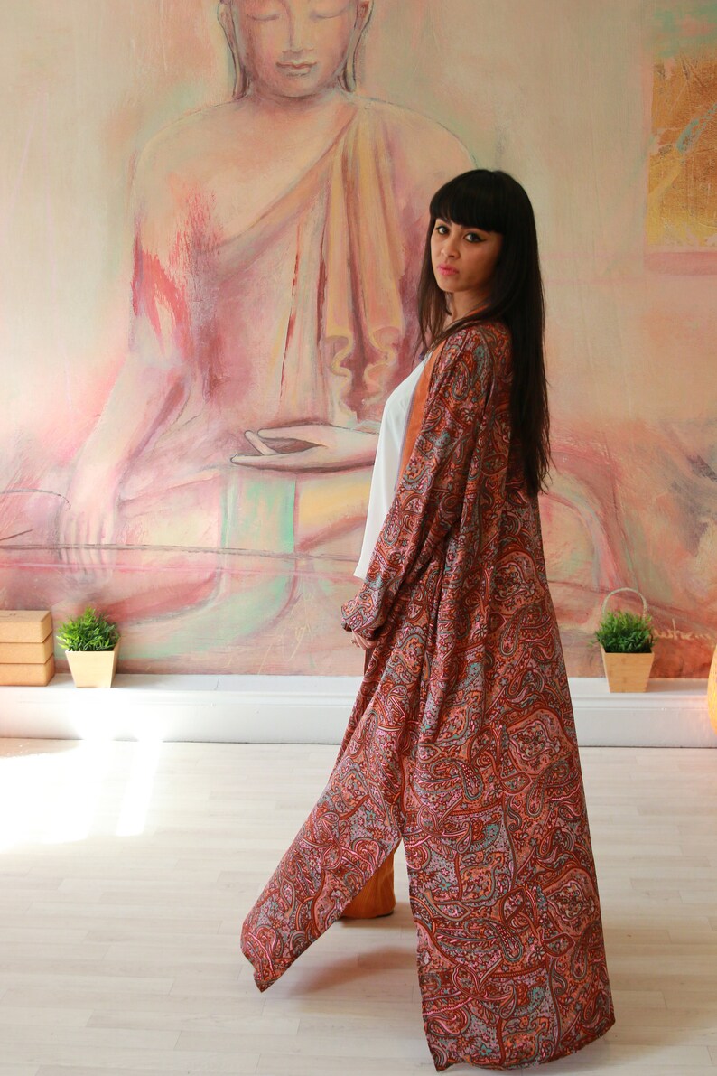 Puede incluir: Una mujer lleva una larga t&uacute;nica de estampado paisley con un top blanco debajo. La t&uacute;nica es una mezcla de colores rojo, naranja y azul. La mujer est&aacute; de pie frente a una pintura de un Buda meditando.