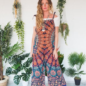 Può includere: Tuta colorata con motivo paisley nei toni dell'arancione, verde acqua e blu navy. La tuta a gamba larga è abbinata a un top bianco con spalle scoperte e stivali con plateau blu. Una lunga collana d'argento completa l'outfit.