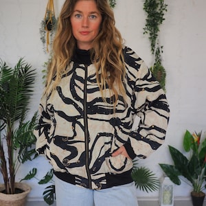 May include: A black and cream-colored bomber jacket with a bold zebra stripe pattern. The jacket has a black collar, cuffs, and hem, and a front zipper closure. The model is wearing light blue jeans.