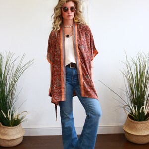 Puede incluir: Una mujer con un kimono de manga larga con estampado, una blusa blanca y pantalones vaqueros azules. Lleva gafas redondas y un collar.