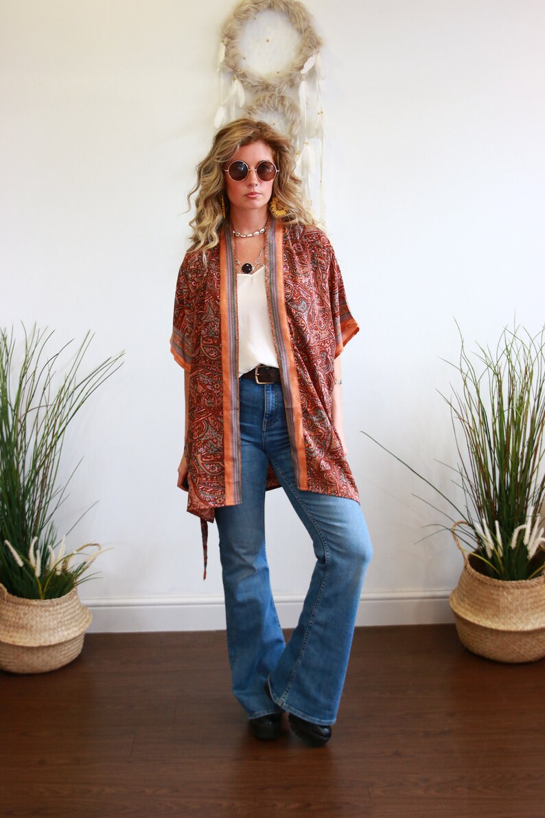 Puede incluir: Una mujer con un kimono de manga larga con estampado, una blusa blanca y pantalones vaqueros azules. Lleva gafas redondas y un collar.