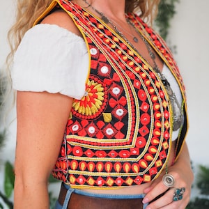 May include: A black cropped vest featuring intricate red and yellow embroidery, small circular mirrors, and yellow trim, worn over a white short-sleeved top with blue denim jeans and a brown leather belt, accented by sunflower earrings.