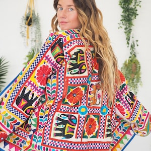 May include: A brightly coloured, embroidered jacket with a pattern of elephants, flowers, and geometric shapes. The jacket has a white base with red, yellow, blue, and green accents. The sleeves and edges are trimmed with a blue border.