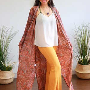 Puede incluir: Una mujer con un kimono largo y fluido con estampado de paisley, un top blanco de tirantes y pantalones amarillos mostaza. Est&aacute; sonriendo y mirando a la c&aacute;mara.