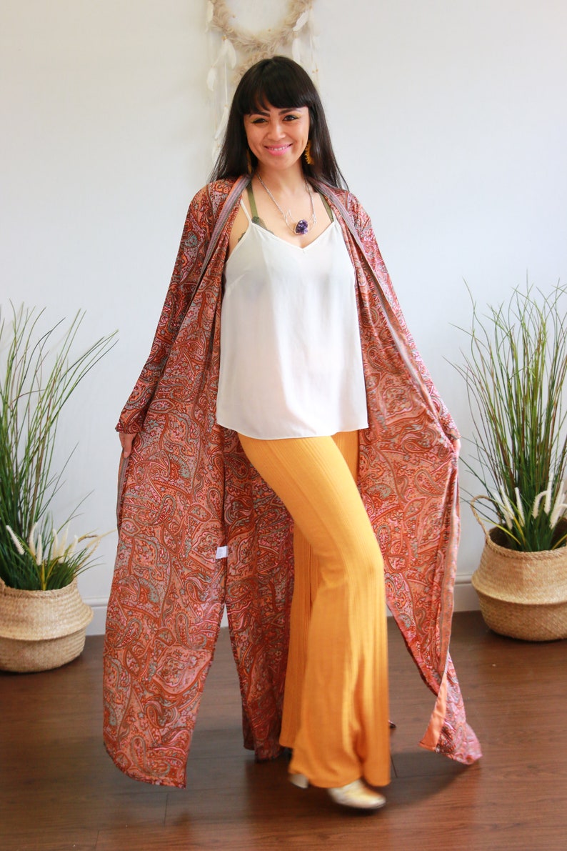 Puede incluir: Una mujer con un kimono largo y fluido con estampado de paisley, un top blanco de tirantes y pantalones amarillos mostaza. Est&aacute; sonriendo y mirando a la c&aacute;mara.