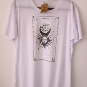 May include: White t-shirt featuring a black and white illustration of a tarot card. The card depicts a sun and moon with a face, and the text "THE TOWER" is printed above the image.