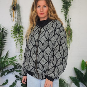 May include: A black and white patterned zip-up jacket with a geometric design. The jacket has a black collar and cuffs. The model is wearing light blue jeans. The background includes hanging plants and other greenery.