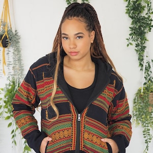 May include: A black zip-up hooded jacket with a vibrant geometric pattern in red, green, and yellow. The jacket features a black lining and is worn over a black top. The model is wearing burgundy trousers.
