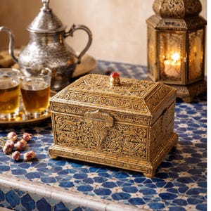 May include: A gold-colored, rectangular box with intricate floral and scrollwork carvings. The box has a hinged lid with a small red gem on top and a decorative clasp. The box is placed on a blue and white tiled surface.