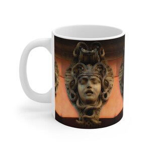 May include: White ceramic coffee mug featuring a detailed image of a Medusa sculpture. The sculpture has a serene face, adorned with snakes and intricate headwear. The background is a warm, reddish-orange hue.