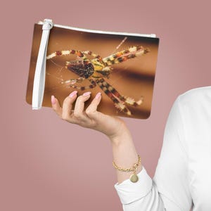 Pode incluir: Uma clutch retangular com fecho de correr e uma alça de pulso branca. A clutch apresenta uma imagem detalhada de uma aranha com um padrão castanho, amarelo e vermelho. O fundo é castanho desfocado.