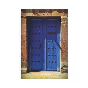 May include: A bright blue wooden door with metal stud detailing, set in a rustic stone frame. The door has a small brass letterbox and a dark handle. The stone frame is a warm, earthy colour, creating a striking contrast.