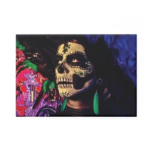 May include: A rectangular canvas print featuring a person with Day of the Dead face paint. The face paint includes a skull design with black and white details, green accents, and long black hair. The background includes colorful floral elements and blue fabric.