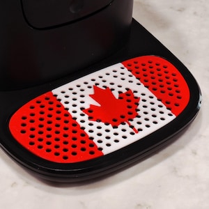 May include: Black coffee machine base with a red and white Canadian flag design. The flag features a red maple leaf on a white background, framed by red perforated sections.