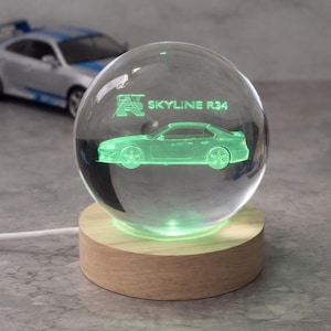 Puede incluir: Una esfera de vidrio transparente con un diseño de coche iluminado en verde y el texto "SKYLINE R34". La esfera se asienta sobre una base de madera con un brillo verde. Un coche de juguete es visible en el fondo.