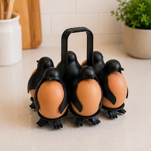 Puede incluir: Un portahuevos negro con forma de pingüino que contiene seis huevos marrones. El soporte tiene un asa para facilitar el transporte. Los huevos están colocados en el cuerpo del pingüino, creando un accesorio de cocina divertido y funcional.