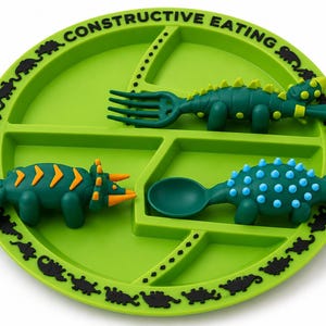 May include: A bright green divided plate with the text CONSTRUCTIVE EATING printed along the rim, featuring a black dinosaur silhouette border. Included are three dinosaur-shaped utensils: a green fork, a green spoon, and a green toy figure.