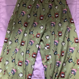 May include: Green pajama trousers with a repeating pattern of cartoon characters wearing antlers, masks, and arrows. The characters have different hair colours, including red, pink, and green. The trousers have an elasticated waistband.