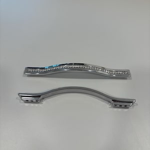 May include: Two silver cabinet handles. One handle is curved with a row of clear rhinestones. The other handle is a simple curved design. Both handles have a polished, reflective surface and are set against a white background.
