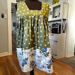 May include: A sleeveless apron featuring an olive green upper section with a white daisy pattern and a gathered waist. The lower section is a light gray fabric decorated with blue roses, green leaves, and black butterfly illustrations.