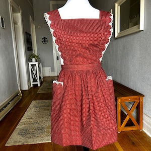 May include: A red polka-dot apron with white lace trim. The apron features a square neckline, a fitted waist, and two front pockets. The edges are trimmed with white lace. The apron is displayed on a mannequin.