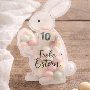 May include: A transparent bunny-shaped container filled with pastel sweets. The container features the number 10 and the words "Frohe Ostern". Several sweets are scattered on the wooden surface.