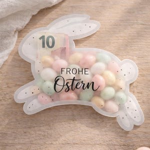 Może przedstawiać: Przezroczysta torba w kształcie królika wypełniona pastelowymi cukierkami. Torba ma numer "10" i napis "FROHE Ostern". Torba leży na drewnianej powierzchni.