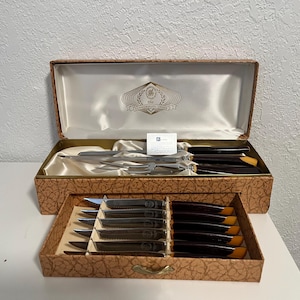 Puede incluir: Un juego de cubiertos vintage en una caja decorativa. El juego incluye cuchillos, tenedores y utensilios para servir con mangos de color marrón oscuro y hojas de acero inoxidable. La caja está forrada con satén y tiene un exterior estampado.