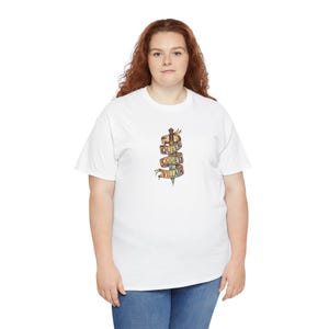 May include: A white short-sleeve t-shirt featuring a central graphic of a silver sword wrapped in a multi-colored ribbon. The ribbon displays the text "QUEER CODED VILLAIN" in bold, dark lettering. The design is positioned on the chest area.