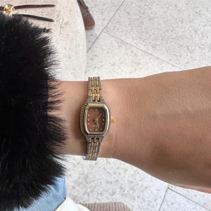 May include: A vintage wristwatch with a rectangular gold-toned face and a brown dial with gold-toned hour markers. The watch has a two-tone silver and gold band. The watch is on a wrist with a black fur sleeve.
