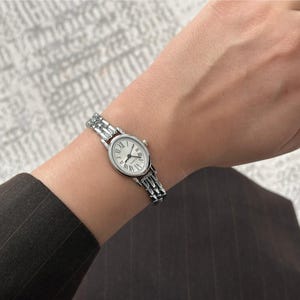 May include: A silver-toned wristwatch with a white face and Roman numeral hour markers. The watch has a linked bracelet and is worn on a wrist. The watch face is oval-shaped and has black hands.
