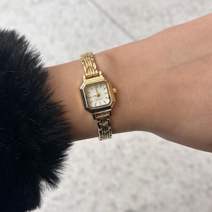 Puede incluir: Un reloj de pulsera dorado con una esfera rectangular y una esfera blanca. El reloj tiene una correa dorada y se lleva en la muñeca. La esfera del reloj tiene agujas y marcadores dorados. Un accesorio clásico y elegante.