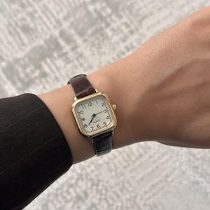 May include: A gold-toned square watch with a white face and black numerals. The watch has a dark brown leather band and is worn on a wrist. The watch face displays the time.