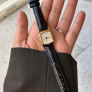 May include: A gold rectangular wristwatch with a cream face and black Roman numeral hour markers. The watch has a black leather strap with a crocodile pattern.