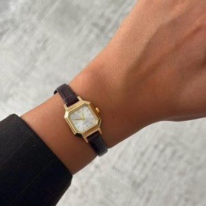 May include: A gold-toned quartz wristwatch featuring an octagonal case and a white dial with simple hour markers. The watch is attached to a dark brown leather strap with a textured, embossed finish that mimics a crocodile skin pattern.