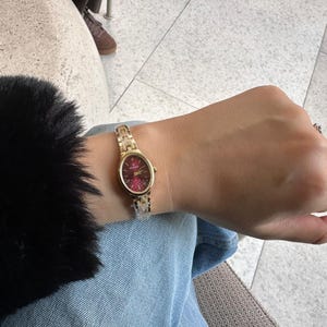Puede incluir: Un reloj de pulsera dorado con esfera burdeos y brazalete de eslabones se lleva en la muñeca. El reloj tiene una esfera redonda. El fondo incluye mezclilla azul y una manga negra y borrosa.