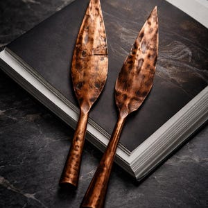 May include: Two copper-coloured serving utensils with leaf-shaped blades rest on a stack of books with a black cover. The utensils have textured handles and a rustic, hammered appearance, suggesting a handmade quality.