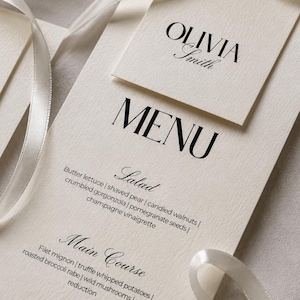May include: A cream-colored paper menu card with black serif and script typography, featuring the name Olivia Smith, tied with a white satin ribbon. The menu lists salad, main course, and dessert options including filet mignon and panna cotta.