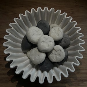 May include: A white ceramic bowl with a ruffled, scalloped edge containing a collection of smooth, round stones. The stones are coloured in shades of white and dark grey, each featuring a small, embossed flower design on the top surface.