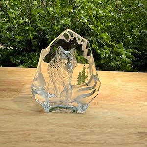 May include: A clear glass sculpture of a wolf, etched with intricate details. The wolf is depicted sitting, with a forest scene in the background. The sculpture has a unique, irregular shape, and is displayed on a wooden surface.
