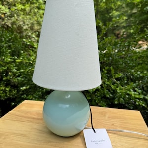 May include: A small, mint-green table lamp with a white lampshade. The lamp has a spherical base and a tapered shade. A black cord and a Kate Spade tag are attached. The lamp is on a wooden surface.