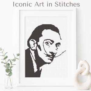 May include: A framed black and white cross-stitch portrait of Salvador Dalí. The artwork is displayed in a white frame and features the text "Iconic Art in Stitches" and "Easy Modern PDF Pattern". A vase with greenery and a decorative white sculpture are in the background.
