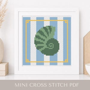 May include: A framed cross-stitch pattern featuring a green seashell design on a blue and white striped background, framed with a gold border. The text "MINI CROSS STITCH PDF" is displayed at the bottom of the frame. The artwork is displayed on a white surface.
