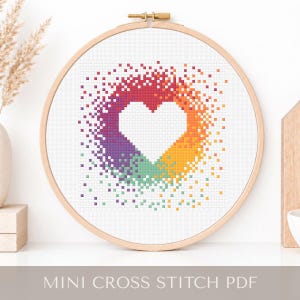 Può includere: Un ricamo a punto croce in un telaio di legno presenta una forma di cuore bianco circondata da una sfumatura arcobaleno pixelata. Il testo "MINI CROSS STITCH PDF" è visualizzato in basso. Il disegno è su tessuto bianco.