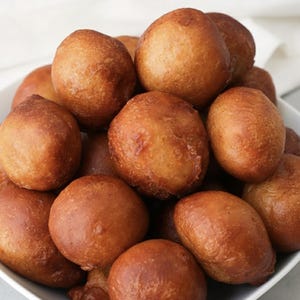 May include: A white bowl filled with numerous golden-brown, spherical fried dough balls. The dough balls have a slightly uneven surface and a rich, warm colour, suggesting they are freshly cooked. The image is a close-up, highlighting the texture and colour of the food.