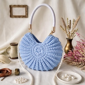 Peut inclure: Un sac à main bleu clair en crochet avec une poignée tressée blanche et des ferrures dorées. Le sac présente un motif circulaire au centre et est orné d'une chaîne de perles. Un article décoratif.
