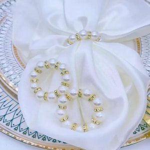 Peut inclure: Deux ronds de serviette en perles blanches avec des détails dorés et des strass transparents. Posés sur une serviette en satin blanc, ils suggèrent une table raffinée. Parfaits pour les occasions spéciales, ils ajoutent une touche sophistiquée.