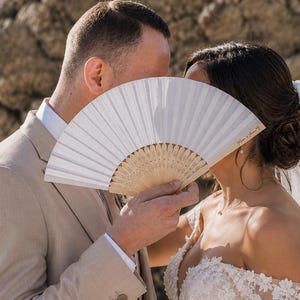 Peut inclure: Un éventail pliant blanc avec une monture en bois clair, portant l'inscription "Ryan & Jenna" en écriture cursive sur le côté droit. L'éventail est tenu devant les visages de deux personnes vêtues de tenues de mariage, dont un costume beige.
